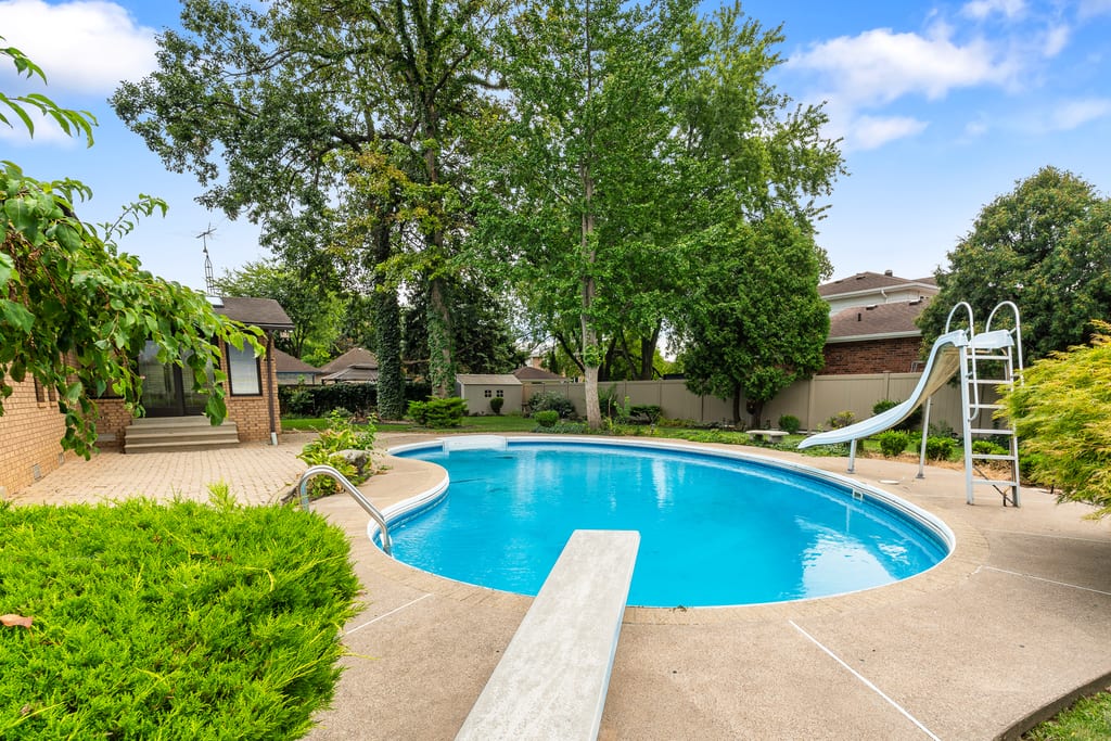 In-ground pool with a diving board and slide, surrounded by lawn and mature trees