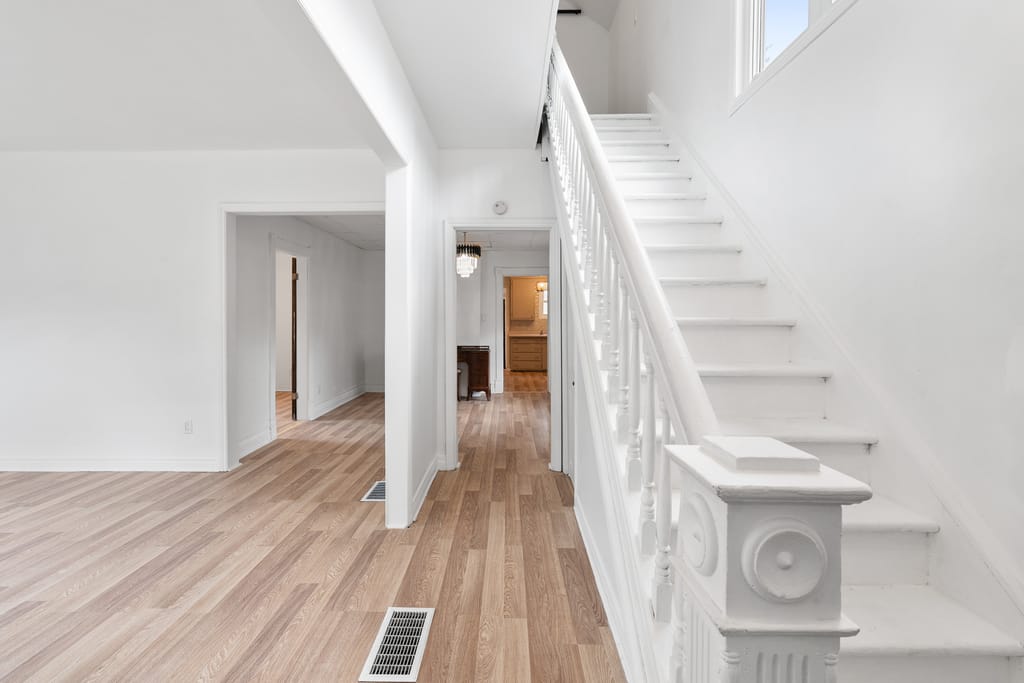 Front hall with a white painted staircase and carved newel post, light plank flooring, and doorways through to the kitchen