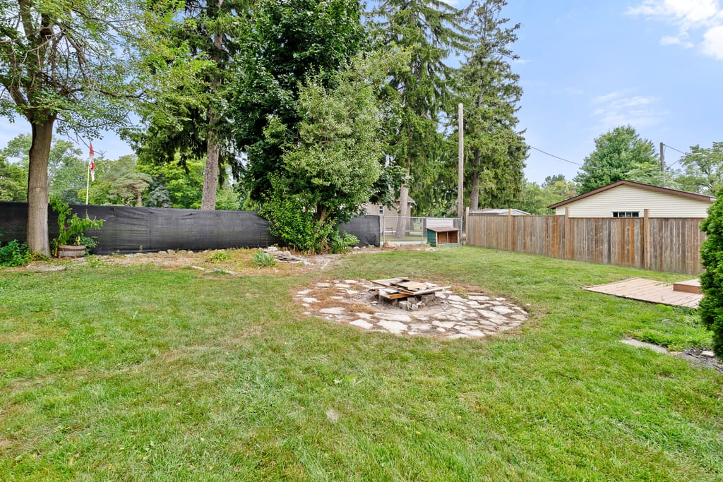 Wide backyard lawn with a stone fire pit circle, tall evergreens, and a wood privacy fence