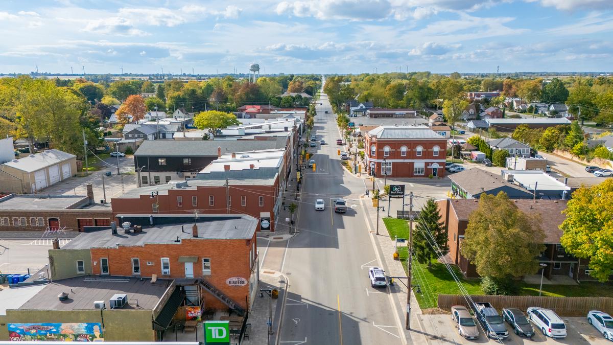 Aerial view over downtown Harrow: brick storefronts along the main street, homes and a water tower beyond, and farmland stretching to the horizon.